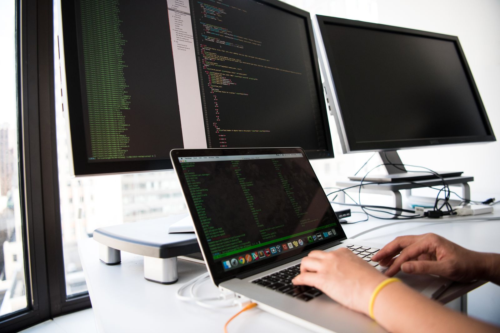 Hands typing on a laptop flanked by monitors showing code